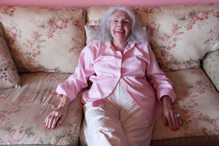 Color photograph of an older woman with gray hair, pink lipstick and nail polish in a pink shirt and white trousers, laughing as she sits back on worn-out sofa with a pink flowery print against a pink wall. 