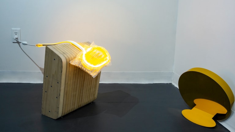 Here is a sculpture made from a layered wood block and yellow LED strip, placed in the corner of a room with white walls.