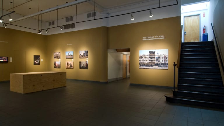 Installation view of “Testimony to War” exhibition showing large wooden rectangle in the middle of the room, photos on the back wall, and a stairwell to the right.