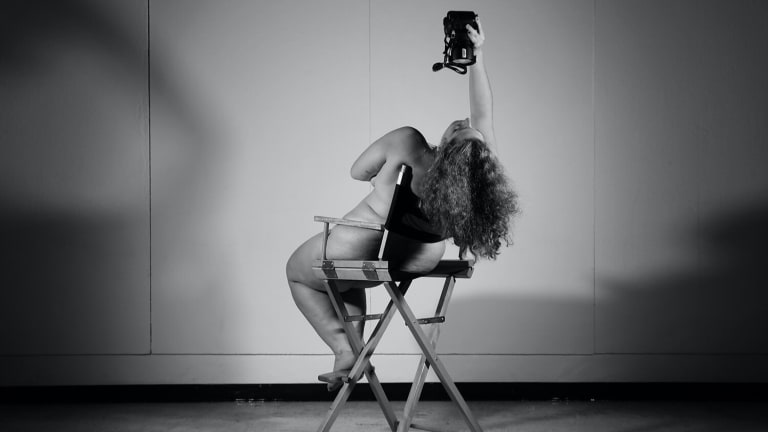 In this black-and-white photo, an artist leans back in a director’s chair to snap a self-portrait from a camera held in the hair. 