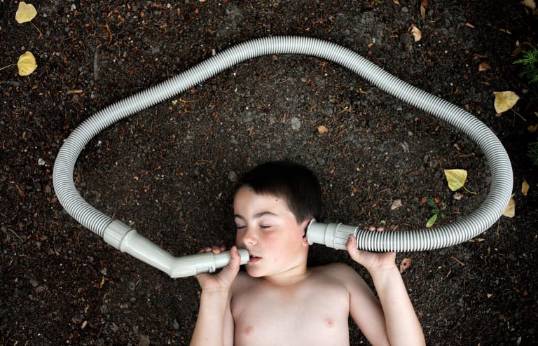 A horizontal color photograph of a shirtless boy lying on the ground with his eyes closed. He is speaking into one end of a vacuum cleaner hose which loops around his head in a cloud shape. He is holding the other end to his ear. 