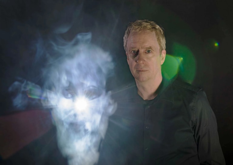 A man in a black shirt standing in front of an illuminated white smoke in the shape of a face