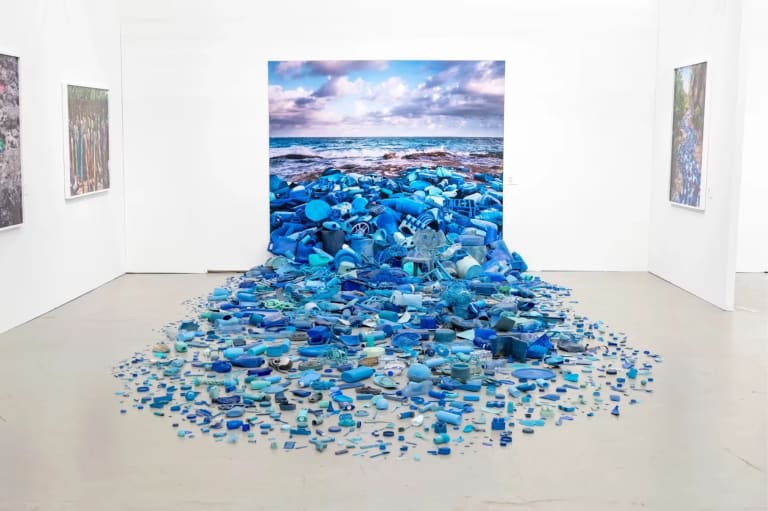 A gallery space with a landscape picture on a wall and blue pieces of plastic spilling out onto the floor.