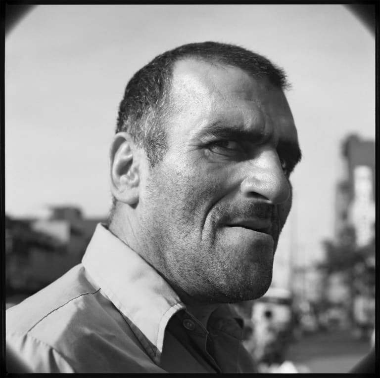 Black and white photographic portrait of a man’s face. He has close-cropped hair and stubble, and is turning toward us with an expression of suspicion.