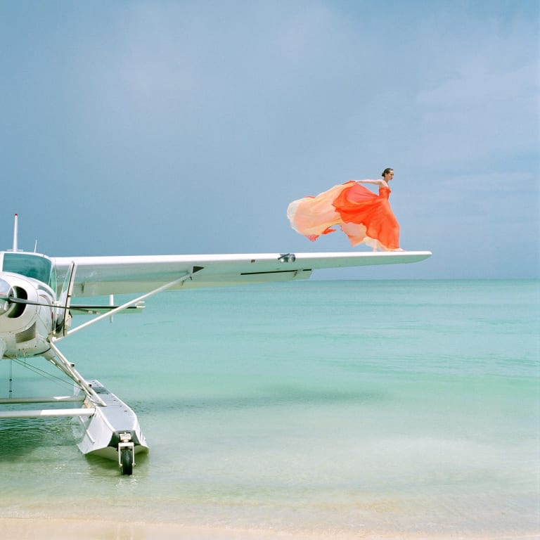 Alt Text: A woman in an orange dress standing on the edge of the wing of a sea plane. She is surrounded by turquoise ocean water and her dress is blowing dramatically in the wind.
