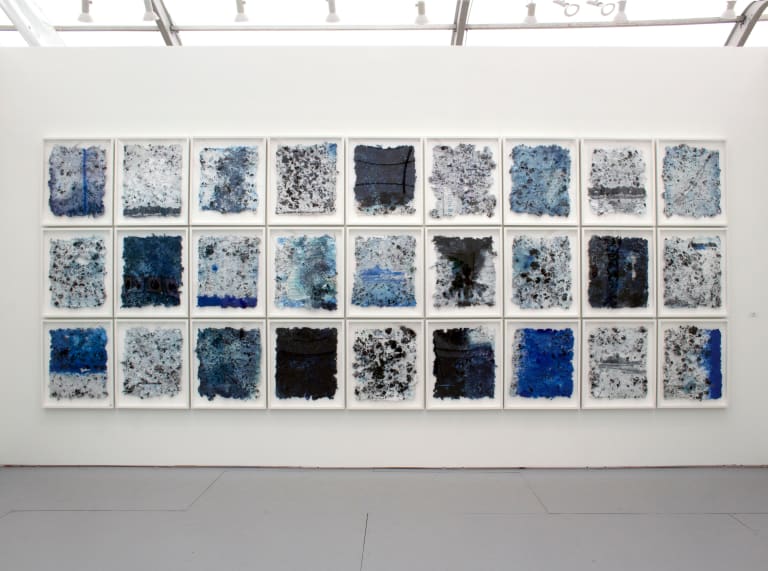 A photograph of a grid of 27 primarily blue artworks hung on a gallery wall.