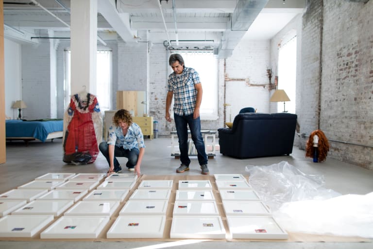 In an open, industrial looking space, a man stands next to a woman who is crouched. They look down at a grid of white frames arranged on the ground.