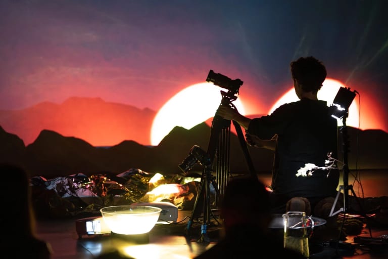 Image of a red sunset over mountains in the background, with a backlit figure operating a camera on a tripod in the foreground.