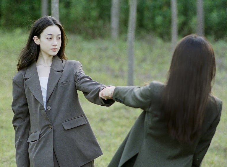 A photo of two women standing in a field in suits, holding hands and leaning away from each other. 