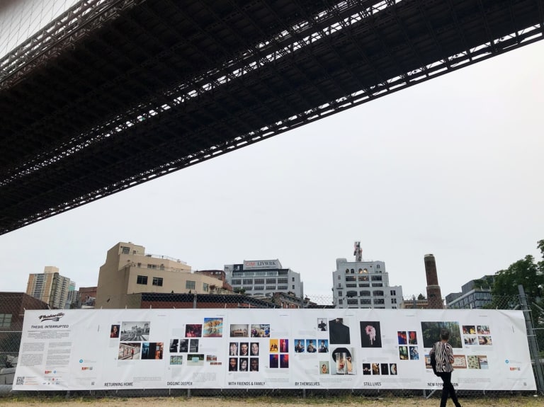 A photograph of an outdoor exhibition of photography in a city park, beneath a large bridge.