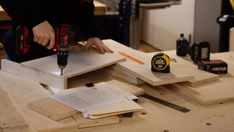Hand using a drill on a piece of wood. Measuring tape, ruler, and other tools on the table.