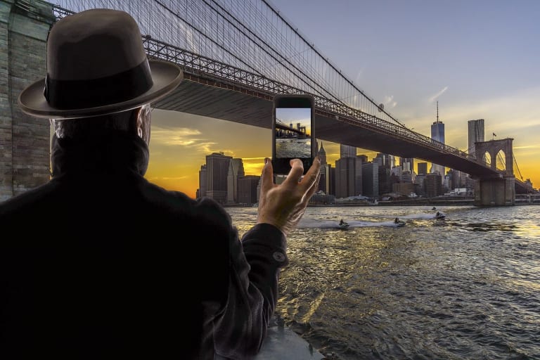 A man, wearing a brown fedora and a black coat, whose back is turned toward us, is looking across the Brooklyn Bridge at the Lower Manhattan skyline against the sunset. His right hand is raised as he is taking a photo of the skyline on his cell phone.