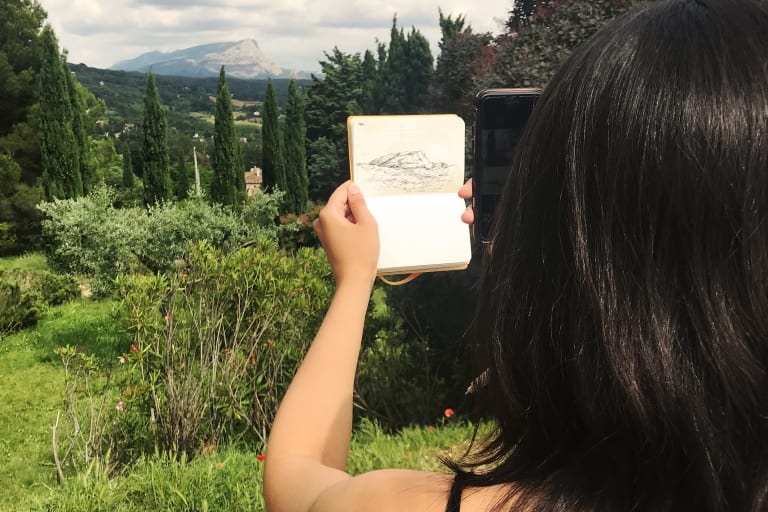 A person holds up a small sketchbook with a drawing of a hill on the page and takes a picture with a very green, hilly scene behind it