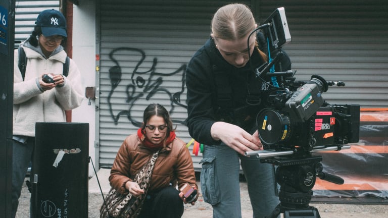 The individuals stand in an outdoor urban setting. The one on the far right is a blonde woman who is wearing a black hoodie and adjusting a large film camera. The other two are working a sound machine and wearing outdoor coats. 