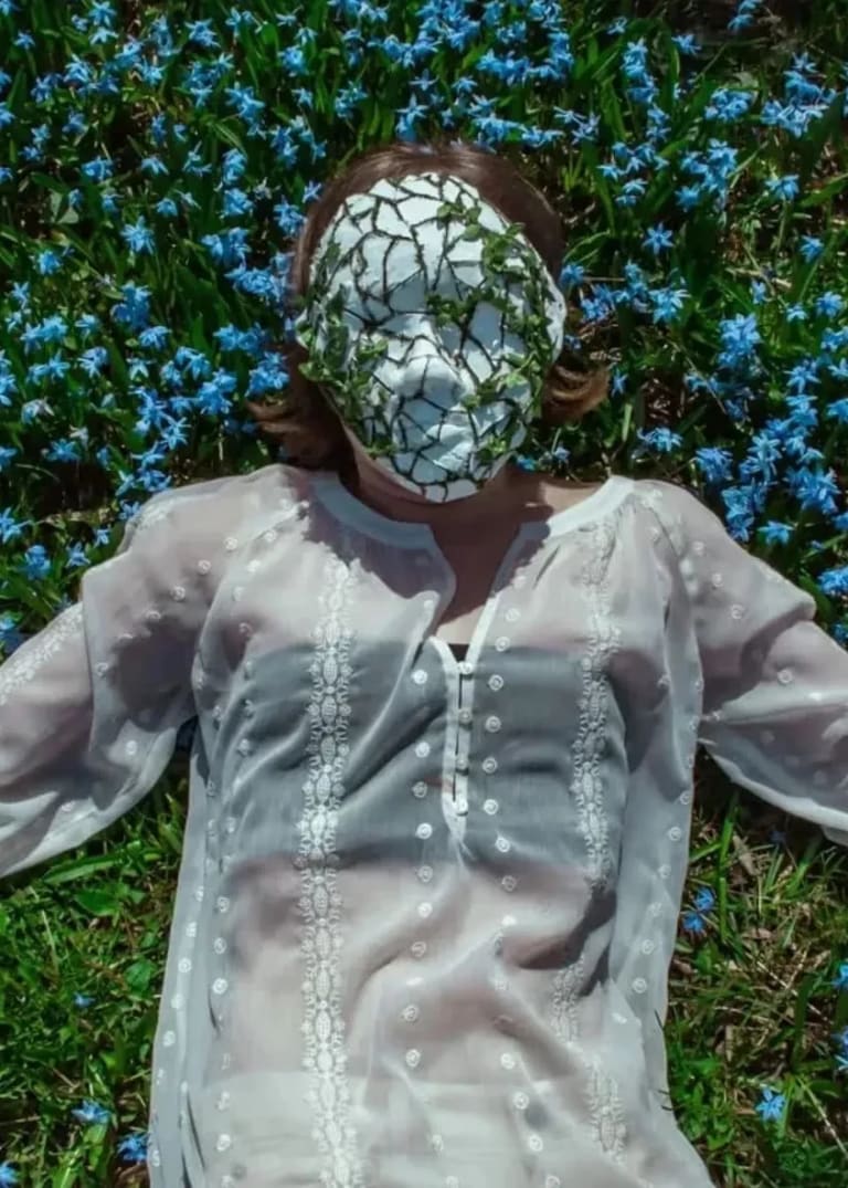 a pale person in a sheer white shirt lies in a field of flowers. There's a mask on their face that's cracked and has leaves growing out of it. 