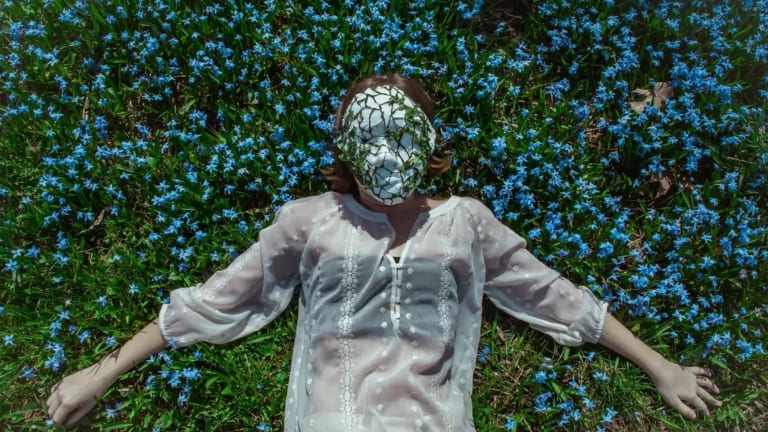 a pale person in a sheer white shirt lies in a field of flowers. There's a mask on their face that's cracked and has leaves growing out of it. 