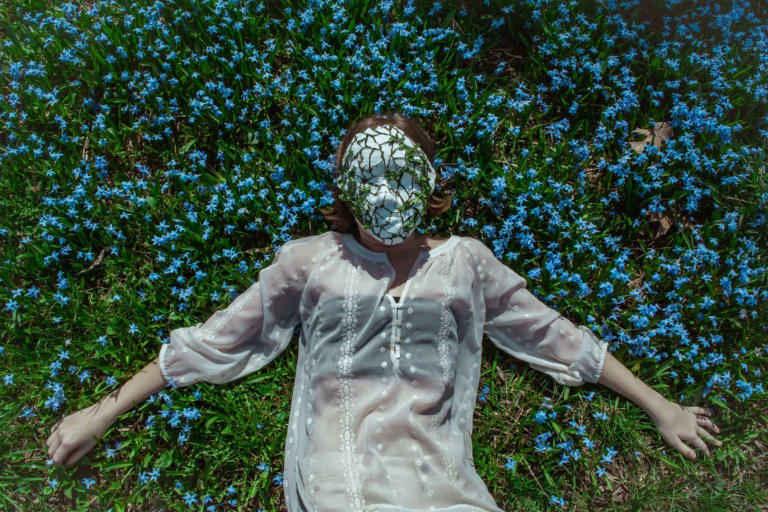 a pale person in a sheer white shirt lies in a field of flowers. There's a mask on their face that's cracked and has leaves growing out of it. 