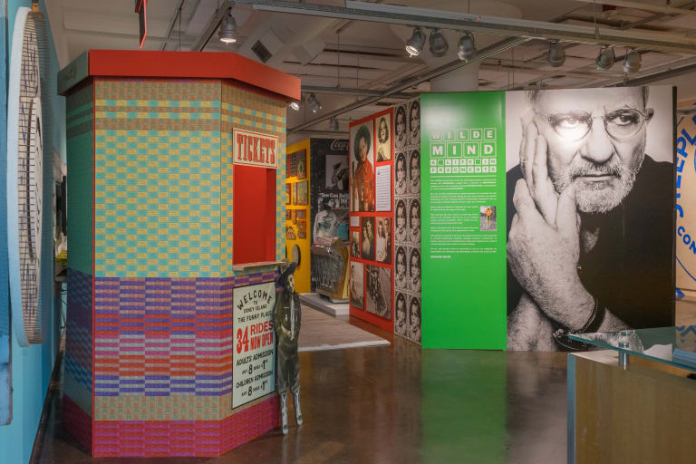 Gallery view of Wilde Mind: A life in fragments, features several walls printed with images and colorful patterns. At the entrance a green wall with white text lists the name of the exhibition and a description of it, next to a large black and white portrait of a man.