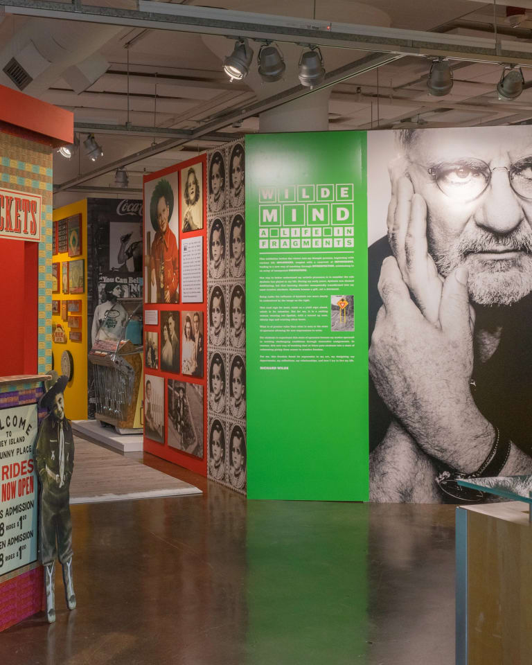 Gallery view of Wilde Mind: A life in fragments, features several walls printed with images and colorful patterns. At the entrance a green wall with white text lists the name of the exhibition and a description of it, next to a large black and white portrait of a man.