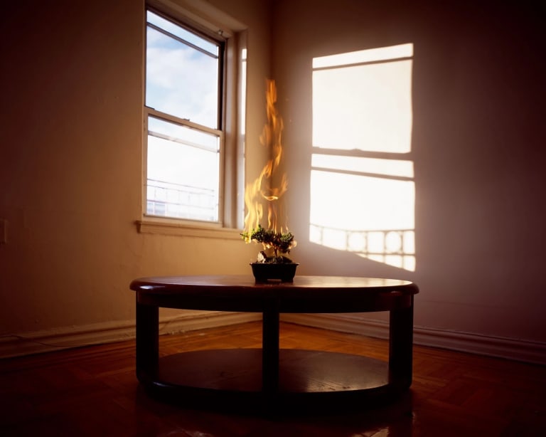 Photograph of a light-filled interior's corner, with a window on the left and a table in the foreground with a large flame atop it.