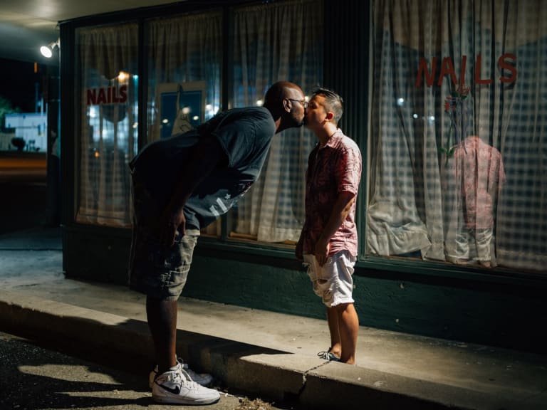 Color photograph of a very tall black man bending over at the waist to kiss a much shorter Asian man on the lips. The setting is the outside of a nail salon at night.