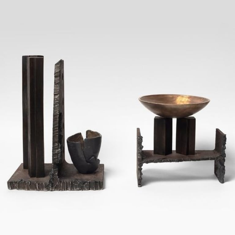 two small metal sculptures, one considting of two upright bars and a small cup on a rectanmgualar base and the other consisting of a gold-colored bowl balanced on two small supports plaved on a table-like platform