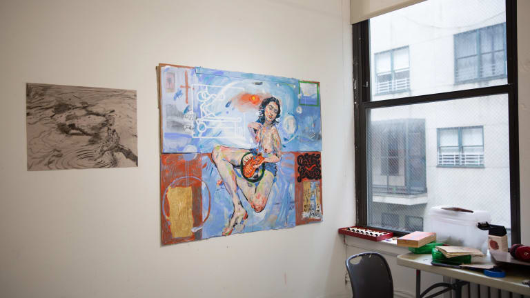 Windowed artist studio in NYC with paintings on the wall and a desk.