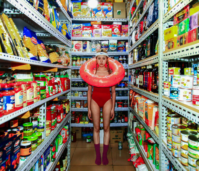 Photograph of a young Asian woman in red one-piece bathing suit, standing in an aisle of a grocery store and staring at the camera, with an inflatable pool ring around her head.