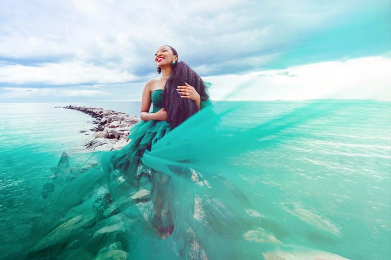 A woman in a teal dress stands smiling on a rock jetty, surrounded by the water.