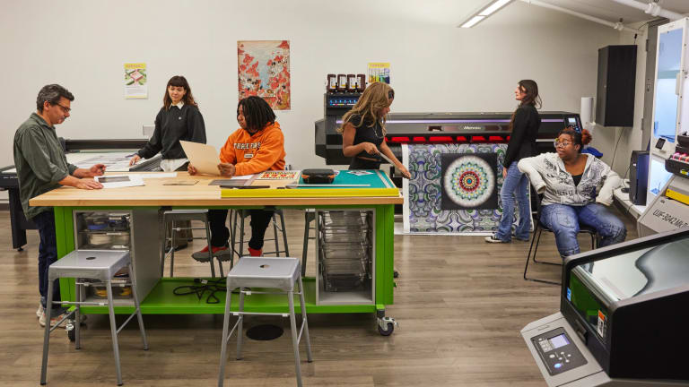 Two separate groups of three people inside of a print shop. One group is having a conversation around a table while the other is around a large print plotter.