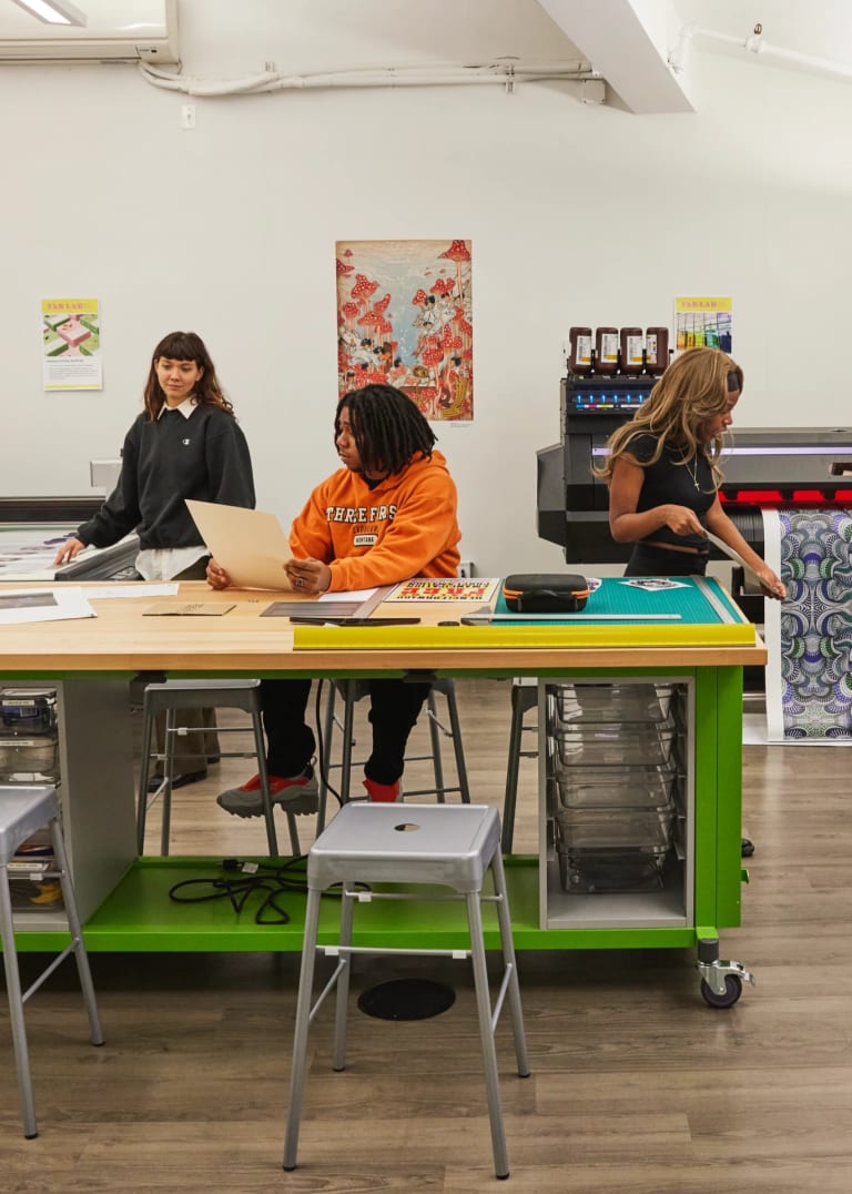 Two separate groups of three people inside of a print shop. One group is having a conversation around a table while the other is around a large print plotter.