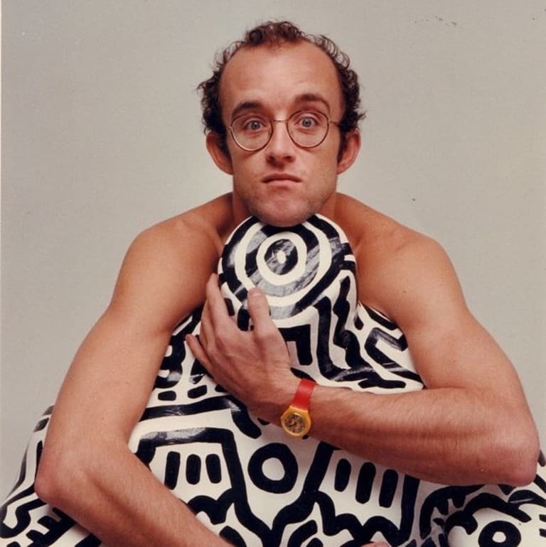 Artist Keith Haring hugs a headless statue from behind, with his head resting on the statue's neck. The statue is white with a black line-based design painted on it.