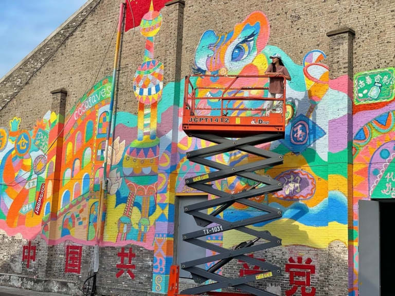 A girl stands on a scissor lift in front of a stone building decorated with a colorful mural.