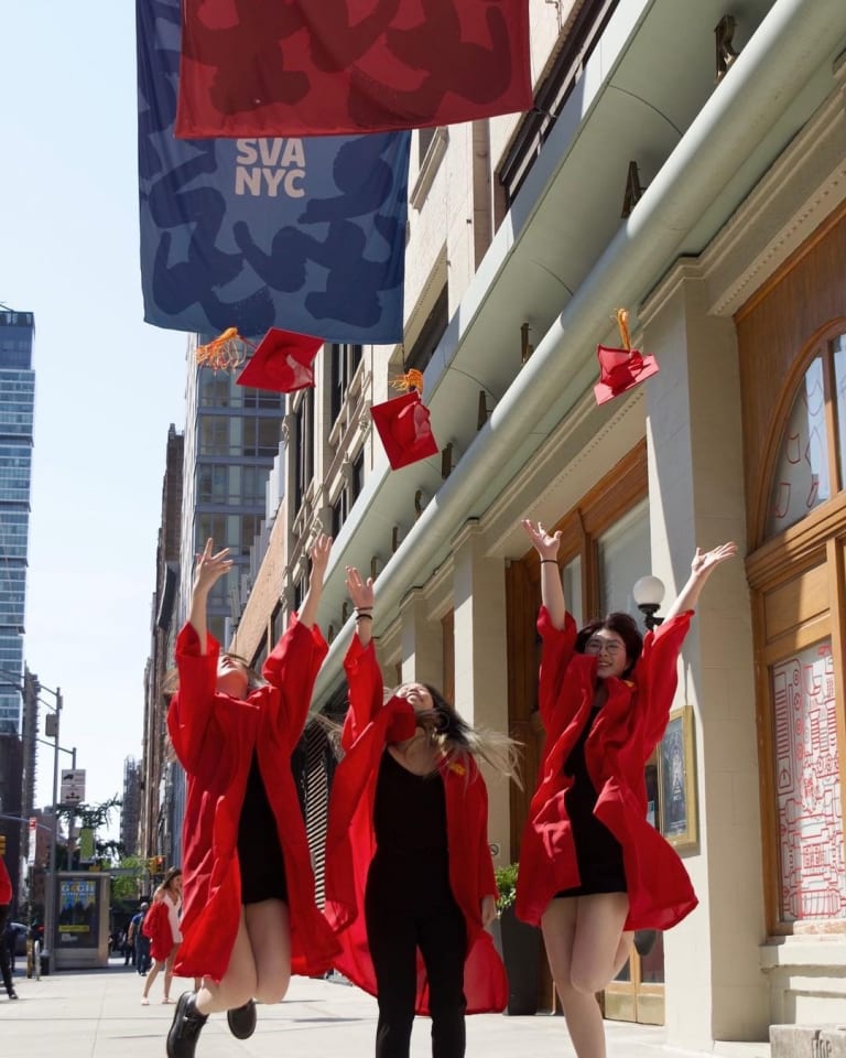 Three members of the SVA class of 2021 throw their red graduation caps in the air, while wearing their red graduation gowns and standing in front of the SVA East 23rd Street location