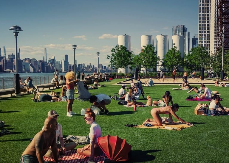 People sit in groups on a grassy area on the water in NYC. It's a clear sunny day and there is a beach volleyball court in the background.