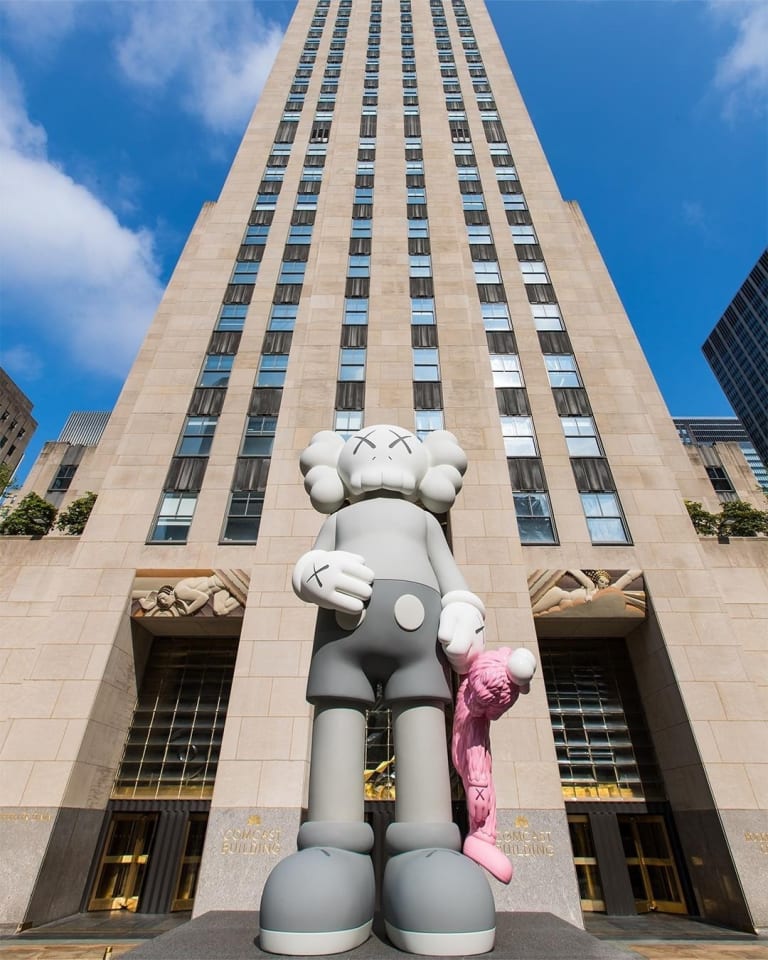 A statue of a Mickey Mouse-esque grayscale character with X-es for eyes holds a pink character at its side. The statue stands tall in front of 30 Rockefeller Plaza in NYC.