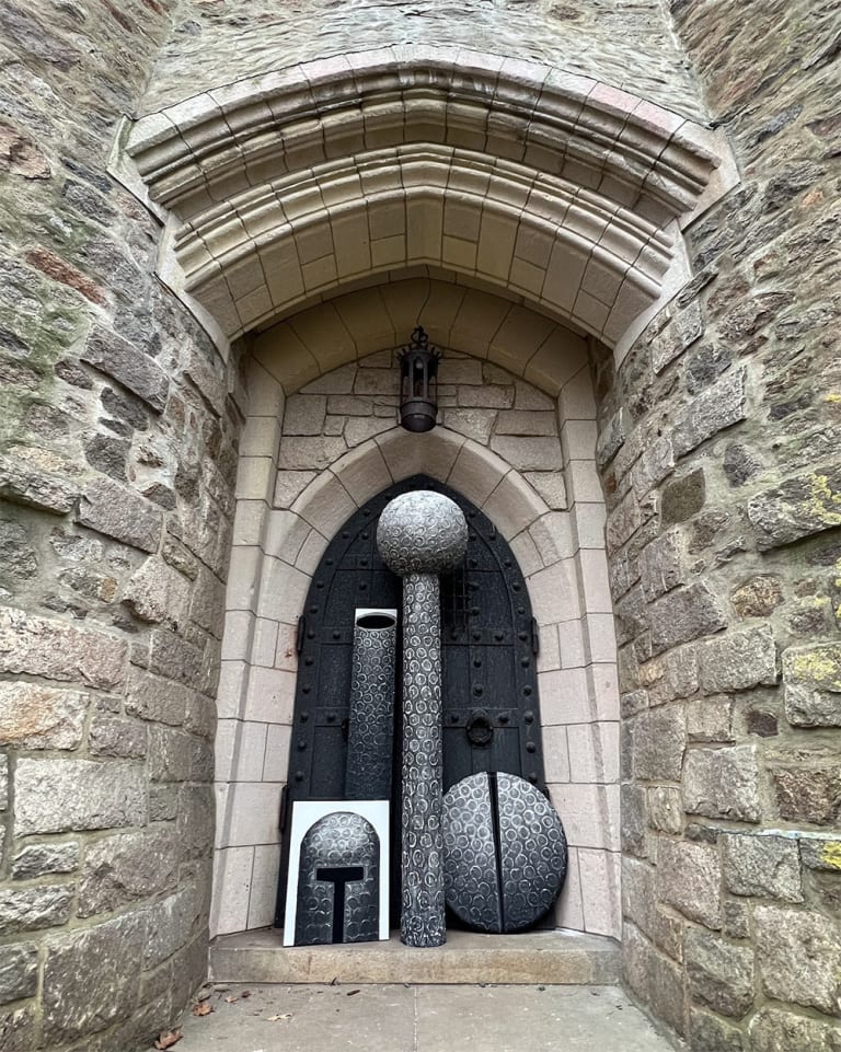 Three paintings, each of a helmet, a shield, and a cylinder stand propped against a castle door. The paintings are accompanied by a painted sculpture in the shape of a cylinder with a sphere on top.