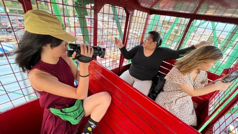An photo of a woman with a camera filming two other people as they sit in a ferris wheel cage