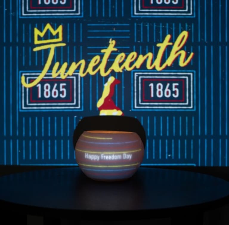 The word Juneteenth (with a crown atop the J) and the year 1865 projected on a wall. In front of the wall is a large bowl shaped object with the words "Happy Freedom Day" projected on it. A torch flame is projected above the bowl shaped object.