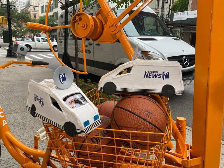 A metal orange shopping cart filled with basketballs and two small models of white Spectrum News NY1 trucks on a New York City sidewalk.