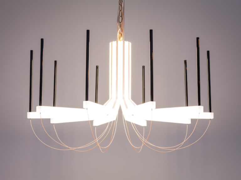 Photo of a chandelier by Eric Forman Studio. There are multiple prongs protruding in U-shapes from the middle. The middle is lit up, and the bottom of the U-shape is a gold wire, and the ends of the U-shapes are coated in black.