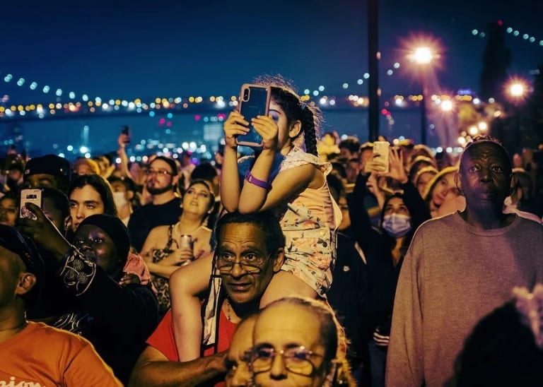 A girl sits atop a man's shoulders with a phone out, presumably recording something. They are in a crowd of people next to the East River.
