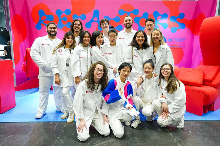 Group of MFA Products of Design students in front of their installation titled "RE-ACTORS," which is printed in a squiggly blue font behind them on bright pink and red fabric.