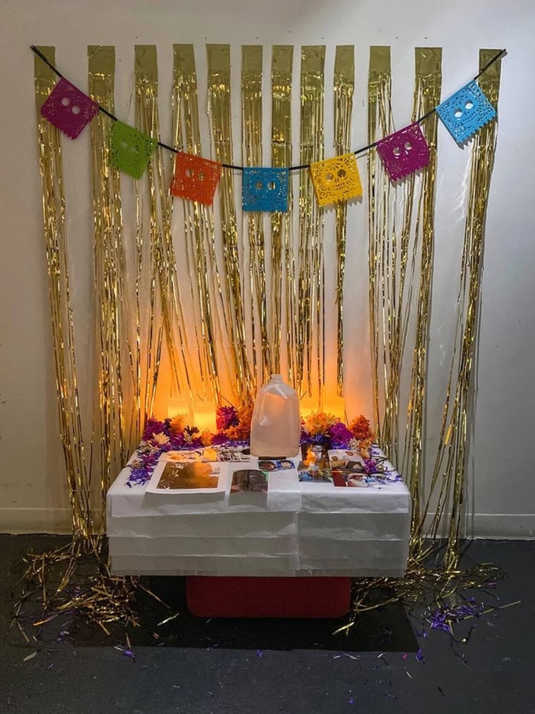 A photo of a low altar next to a white wall with gold fringe hanging on it and colorful flags with sugar skull motifs. On the altar are flowers, a water jug and various photos.