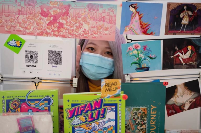 A girl with long purple hair sits behind a booth of drawings and zines. She is wearing a blue mask and holding up her fingers in a peace sign. 