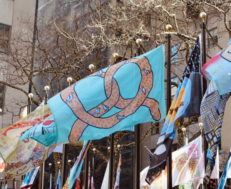 A photo of a pretzel covered with pigeons on a blue flag flying in Rockefeller Center among other flags