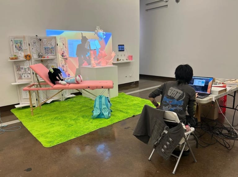 A photo of students installing an art show in a white gallery. The installation includes a stuffed animal character reclining on a pink chair with a projection on the wall in the background.