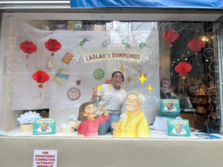 A photo of a person in the window of a book store surrounded by an installation for a book titled "Laolao's Dumplings"