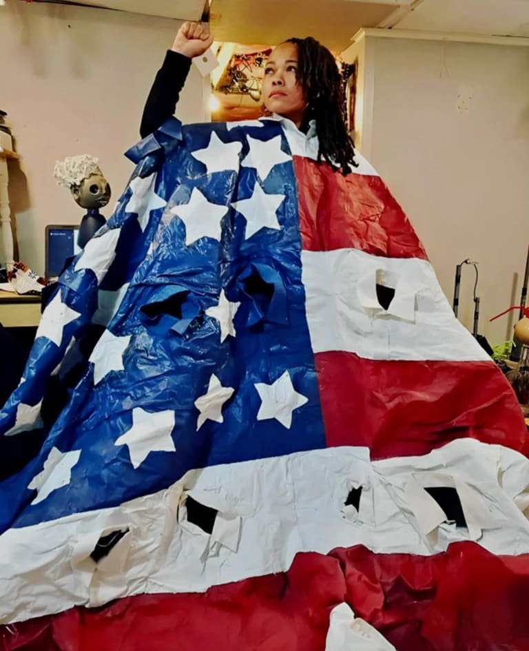 A woman stands engulfed in a plastic American flag with holes in it raising her fist