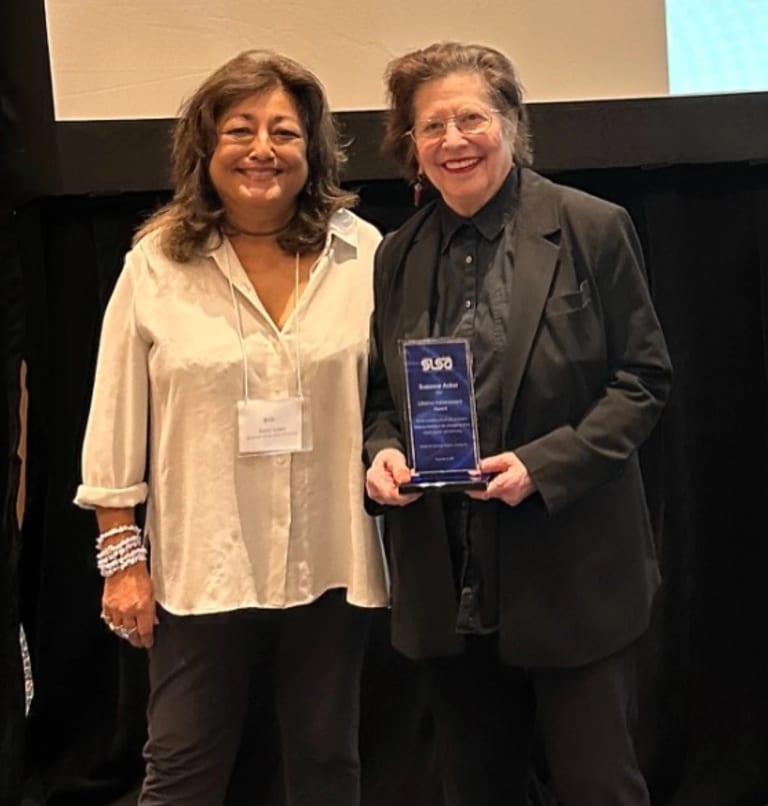 Two women stand next to each other on stage, the woman on the right is holding an award